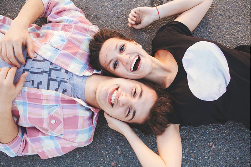 Young couple laying on their backs on pavement looking up at camera with smiles.